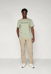 Lys grøn kortærmet t-shirt med sort "Columbia" logo, parret med beige cargo bukser og hvide træningssko, accessoriseret med hvide solbriller.
