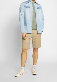 Camicia di jeans di colore azzurro chiaro con due tasche al petto; t-shirt verde oliva sotto, pantaloni beige e sneakers alte bianche.