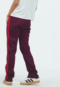 Bordeauxrote Jogginghosen mit roten Seitenstreifen, entspannter Schnitt und elastischem Bund, kombiniert mit weißen Sneakers und schwarzen Streifen.