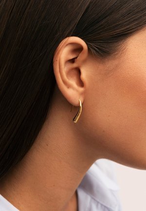 Primer plano de la oreja de una mujer con el cabello oscuro y liso, que lleva un pequeño pendiente dorado, curvado y brillante, sobre un fondo claro.