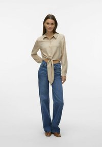Camicia beige con bottoni e dettaglio legato davanti, abbinata a jeans blu a vita alta e gamba larga e scarpe marroni. Tessuto dalla texture liscia.