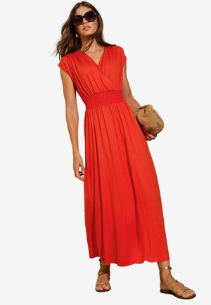Abito lungo senza maniche rosso con vita arricciata, gonna fluente e texture morbida. Accessoriato con una borsa intrecciata e sandali con lacci.