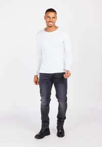 Camicia bianca a maniche lunghe, jeans neri slim-fit e stivali neri allacciati. Il modello sta in piedi con un leggero sorriso su uno sfondo neutro.
