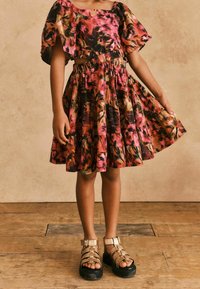 Farbenfrohes, blumengeprägtes Outfit mit einem verkürzten Oberteil und einem gestuften Rock. Der Stoff scheint leicht zu sein. Kombiniert mit klobigen, goldenen Sandalen.