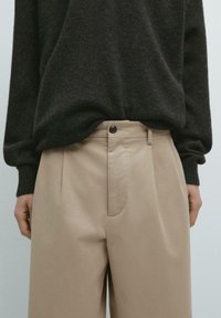 Pantaloni beige su misura con una finitura liscia, caratterizzati da una chiusura con bottone, pieghe e una vestibilità comoda, abbinati a un maglione scuro in maglia.