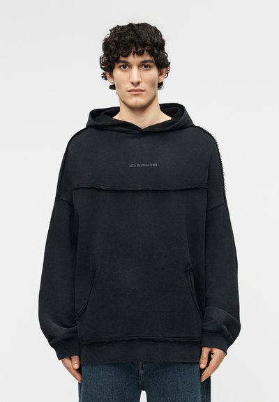 Junger Mann mit lockigem Haar, der einen schwarzen übergroßen Hoodie mit dem Schriftzug "LES BENJAMINS" und blaue Jeans trägt, vor einem schlichten Hintergrund steht.