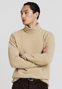 Maglione beige con collo a dolce vita a trama e a costine e maniche lunghe. Modella con le braccia incrociate, indossa pantaloni scuri e una cintura nera.