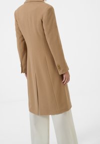 Abrigo camel de corte estructurado, con cuello con solapa, detalles de botones dorados en las mangas y apertura trasera. Combinado con pantalones claros.