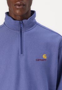 Purple zip-up sweatshirt with a high collar, featuring a Carhartt logo in red and yellow embroidery on the left chest area. Soft material.