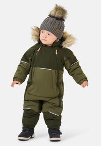 Grüner, isolierter Schneeanzug mit reflektierenden Akzenten, Reißverschlusstaschen und einer mit Fell besetzten Kapuze. Getragen mit einer grauen Strickmütze und schwarzen Stiefeln.