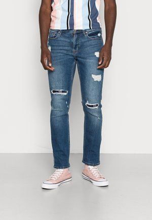 Mand iført slidte blå jeans og rosa høje sneakers, stående på en ensfarvet grå gulv mod en hvid væg.