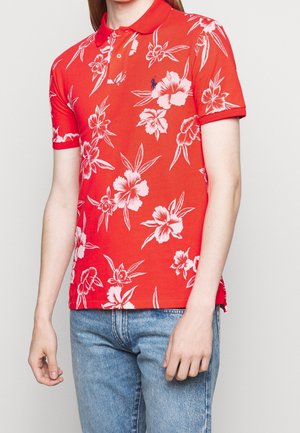 Mężczyzna w czerwonej koszulce polo z krótkim rękawem, z białym kwiatowym wzorem i niebieskim haftowanym logo, w połączeniu z jasnoniebieskimi dżinsami, na jednolitym tle.