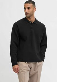 Férfi fekete hosszú ujjú pólót és bézs nadrágot visel, az egyik kezét a zsebébe dugva áll, oldalt nézve egy sima háttér előtt.