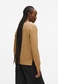 Barna kötött pulóver bordás mandzsettákkal és alsó szegéllyel. Hátul egy kicsit hosszabbra van vágva. Fekete nadrággal párosítva. Hosszú fonott haj látható.