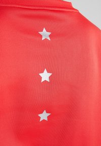 Jersey deportivo rojo con un acabado texturizado que presenta tres estrellas plateadas verticales centradas en la parte posterior. Diseño liviano de mangas cortas.