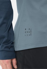 Camicia a maniche lunghe con una manica blu e un corpo azzurro chiaro. Presenta cuciture a vista e un'etichetta stampata nera che recita "RICICLATO ALL'INTERNO".