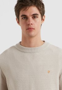 Hellbeige, strukturiertes Strickpullover mit einem gerippten Kragen und einem orangefarbenen Logo-Akzent auf der linken Brust. Gemütlicher, lockerer Schnitt.