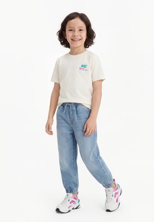 Enfant aux cheveux bouclés foncés souriant, portant un t-shirt blanc Nike, un jean jogger bleu et des baskets blanches avec des accents roses et turquoise.