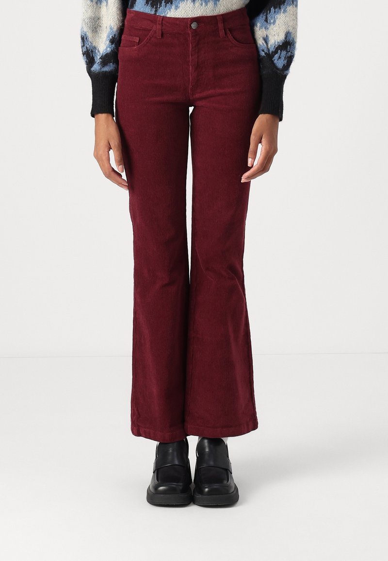Femme portant un pantalon évasé en velours côtelé bordeaux taille haute, associé à des chaussures noires épaisses et un pull à motifs bleus, debout devant un fond uni.