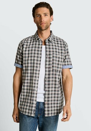 Homme portant une chemise à manches courtes à carreaux retroussées sur un débardeur blanc avec un jean bleu, debout devant un fond clair uni.