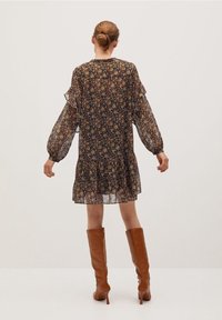 Femme avec les cheveux en chignon portant une robe longue à manches longues à fleurs sombres et des bottes à talons brunes jusqu'aux genoux, se tenant devant un fond uni.