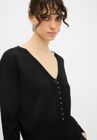 Maglione nero a maniche lunghe con scollo a V, caratterizzato da una fila di bottoni decorativi, un tessuto lavorato e una silhouette aderente.