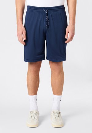 Mężczyzna ubrany w granatowe siateczkowe szorty z białym sznurkiem, białe skarpetki do kostek oraz kremowe trampki, stojący na jednolitym tle.