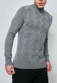 Pull en tricot gris présentant un motif en cable, un col et des poignets côtelés, avec une coupe ajustée mettant en valeur le tissu texturé.