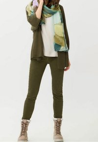 Cardigan de canalé en verde oliva sobre una blusa blanca, combinado con pantalones ajustados en verde oliva y botas beige con borde de piel sintética. Bufanda con estampado en azul claro y verde.