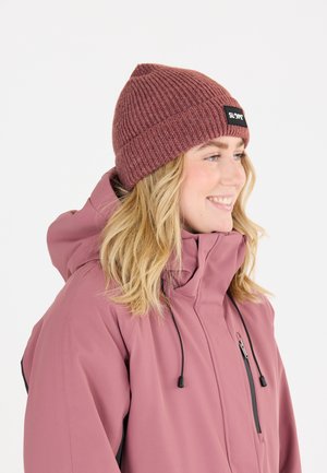Gebreide bordeauxrode beanie met een label aan de voorkant, gedragen met een roze jas met hoge kraag en verstelbare trekkoorden, van gestructureerde stof.