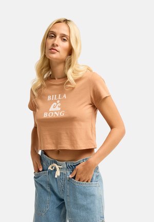 Blondynka w krótkim brązowym t-shircie Billabong i jasnoniebieskich dżinsach ze ściągaczem w pasie, ręce w kieszeniach, neutralne tło.