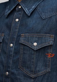 Κοντινό πλάνο σκούρου μπλε τζιν πουκάμισου με μεταλλικά κουμπιά snap, εμπρόσθιες τσέπες με καπάκι, πορτοκαλί ραφές και μικρό πορτοκαλί κεντημένο λογότυπο στην τσέπη.