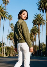 Fiatal nő zöld dzsekiben és fehér nadrágban, aki naplementekor egy pálmafákkal szegélyezett utcán hátrafordulva néz.