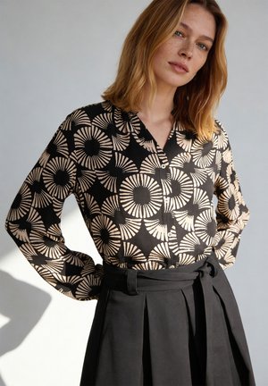 Kobieta w czarnej bluzce z beżowymi okrągłymi wzorami, zestawiona z czarną plisowaną spódnicą z wysokim stanem, stojąca na jednolitym jasnym tle.