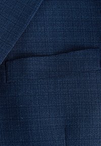 Tissu texturé bleu marine avec un motif rayé subtil. Comprend un détail de poche passepoilée, ajoutant de la structure au design.