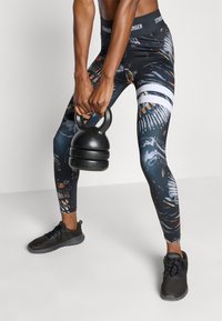 Μαύρο κολάν με σχέδια και πολύχρωμα γραφικά, με ψηλή μέση και εφαρμοστό σχέδιο, συνδυασμένο με μαύρα αθλητικά παπούτσια και ένα κετλμπελ.