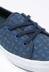 Marinblå kanvask sneaker med ett texturerat mönster, med platta marinblå snören, runda öglor och en vit gummisula.