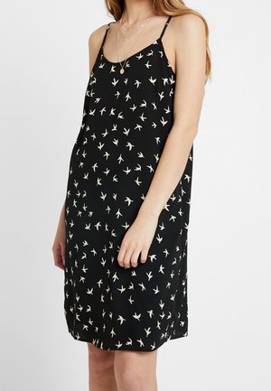 Femme portant une robe noire sans manches avec des motifs d'oiseaux blancs et de fines bretelles, accessoirisée de colliers dorés superposés.