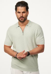 Hellgrünes Strick-Polohemd mit kurzen Ärmeln, halbem Reißverschluss und strukturierter Musterung. Verfügt über einen Kragen und kontrastierende Ärmelausschnitte.