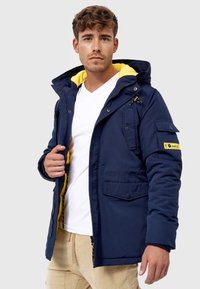 Veste isolante bleu marine avec doublure jaune, poches avant et capuche amovible. Dotée de zippers et d'accents en boutons.