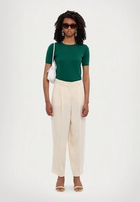 Green short-sleeve top, pleated cream trousers, gold sandals, silver handbag, large sunglasses, textured wavy hair. Simple yet modern ensemble.