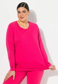Rosa Langarmshirt mit Rundhalsausschnitt, aus weichem Material gefertigt. Das Modell hält den Saum und zeigt eine entspannte Passform und geschmeidige Textur.