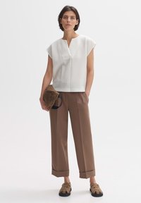 Witte top met patroon en korte mouwen, gecombineerd met bruine wijde broek en bruine sandalen. Houdt een bruine gewatteerde tas vast.