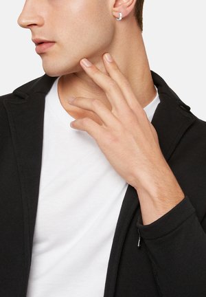 Black zippered jacket with a smooth texture, worn over a white t-shirt. A silver hoop earring is visible in the model's ear.