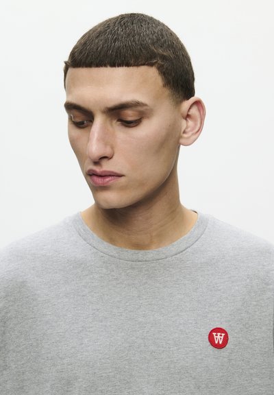 Sweat-shirt gris à col rond en tissu doux avec des manches courtes, arborant un petit logo circulaire rouge sur le côté gauche de la poitrine.