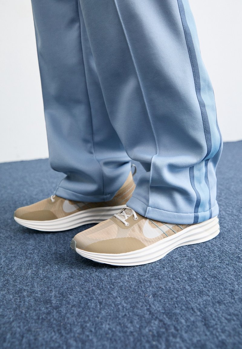 Baskets beige et blanches portées avec un pantalon de survêtement bleu clair ample sur un sol moquetté bleu.
