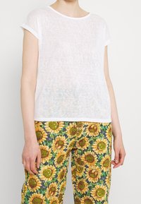 Top corto de manga corta blanca con un tejido texturizado y transparente, combinado con pantalones verdes que presentan un estampado de girasoles en amarillo y marrón.