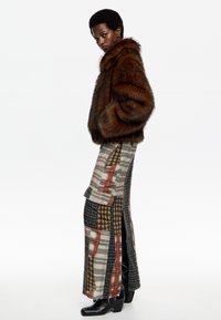 Chaqueta de piel sintética marrón y una falda maxi de cuadros con un patchwork de varios colores y patrones. Botas de tacón negras completan el conjunto.