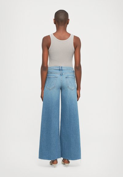 Person standing facing away wearing a beige tank top and high-waisted wide-leg blue jeans with frayed hems and heeled sandals.