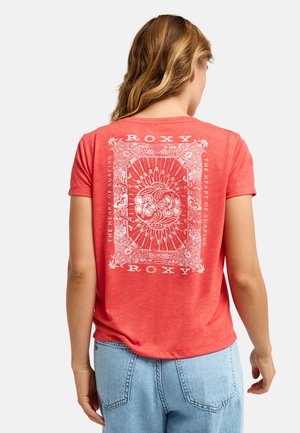 Femme aux cheveux châtain clair portant un t-shirt rouge Roxy avec un motif floral blanc et un texte au dos, ainsi qu'un jean bleu clair.